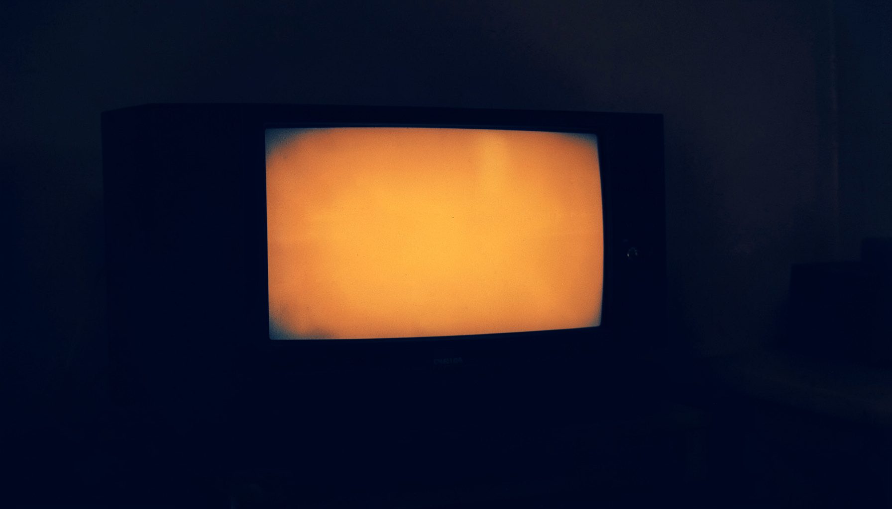 A dark duotone photograph of a television screen glowing in a dim room, deep navy and muted amber tones, moody and cinematic atmosphere.
