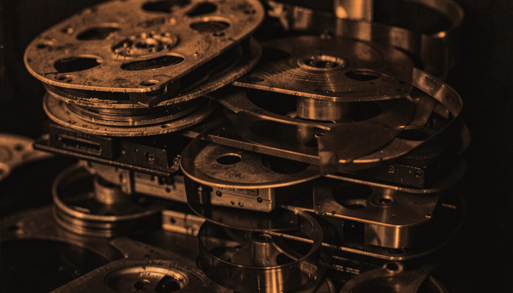 A dark duotone photograph of stacked film reels and tape cassettes, charcoal and warm copper tones, archival atmosphere.