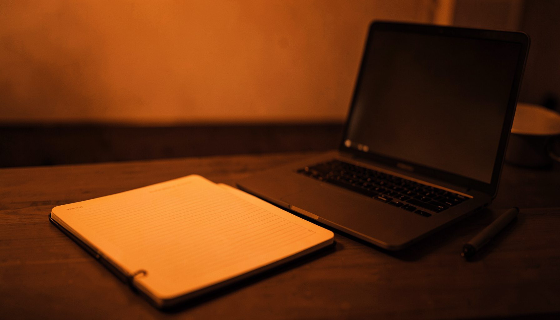 Open notebook and pen on a desk beside a glowing laptop screen, warm amber and deep brown duotone