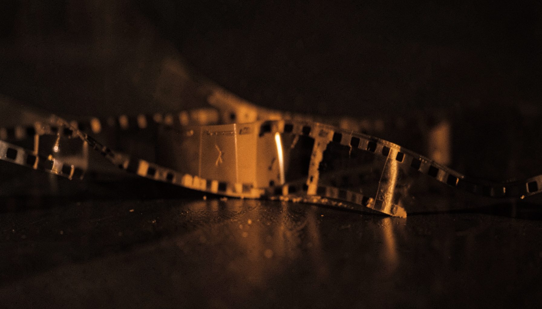 Film strip unspooling across a dark surface with soft amber light catching the edges, warm duotone