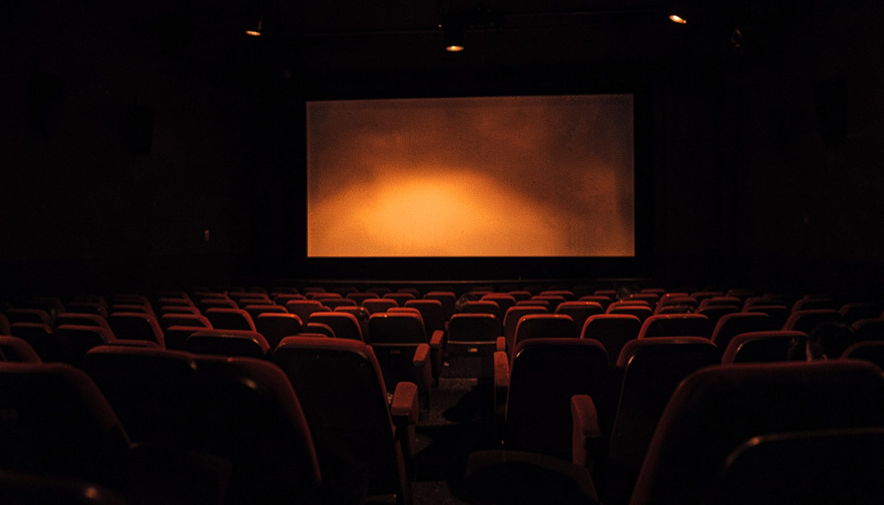 Darkened cinema interior with rows of red seats and a glowing screen, deep shadows and warm amber highlights in a two-tone duotone