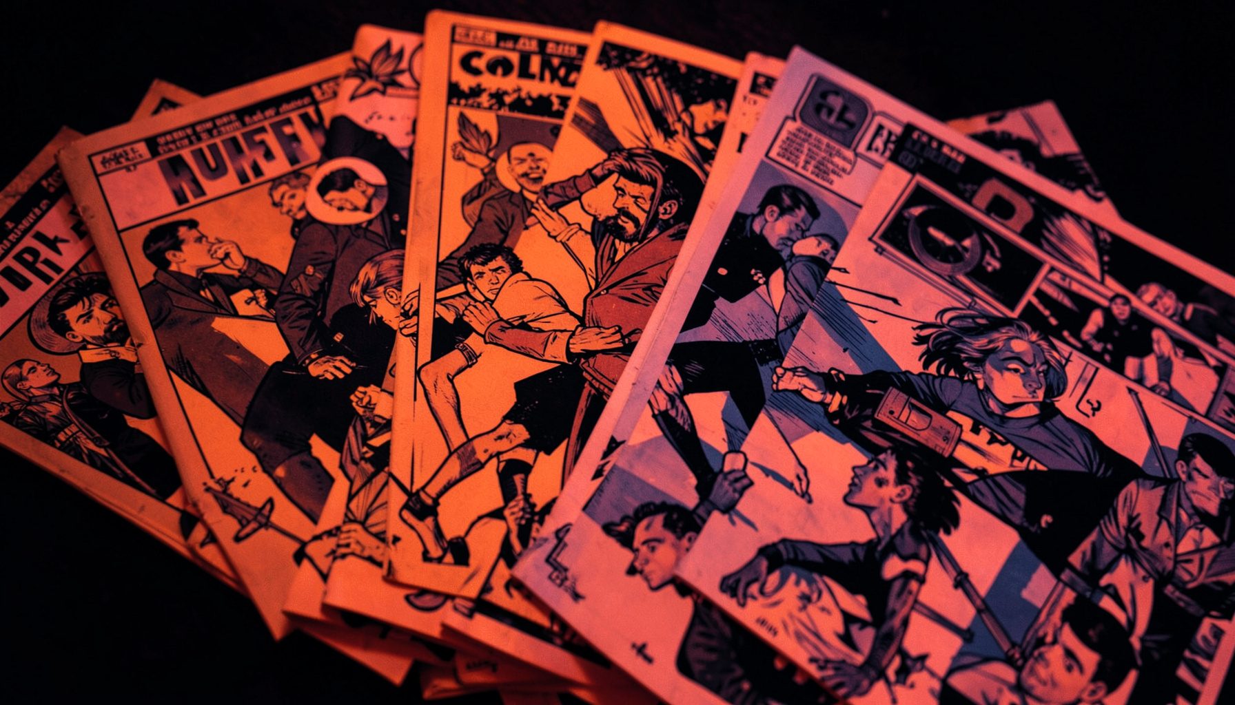 A duotone close-up of colorful comic book covers fanned out on a dark surface, rich crimson and deep indigo tones.