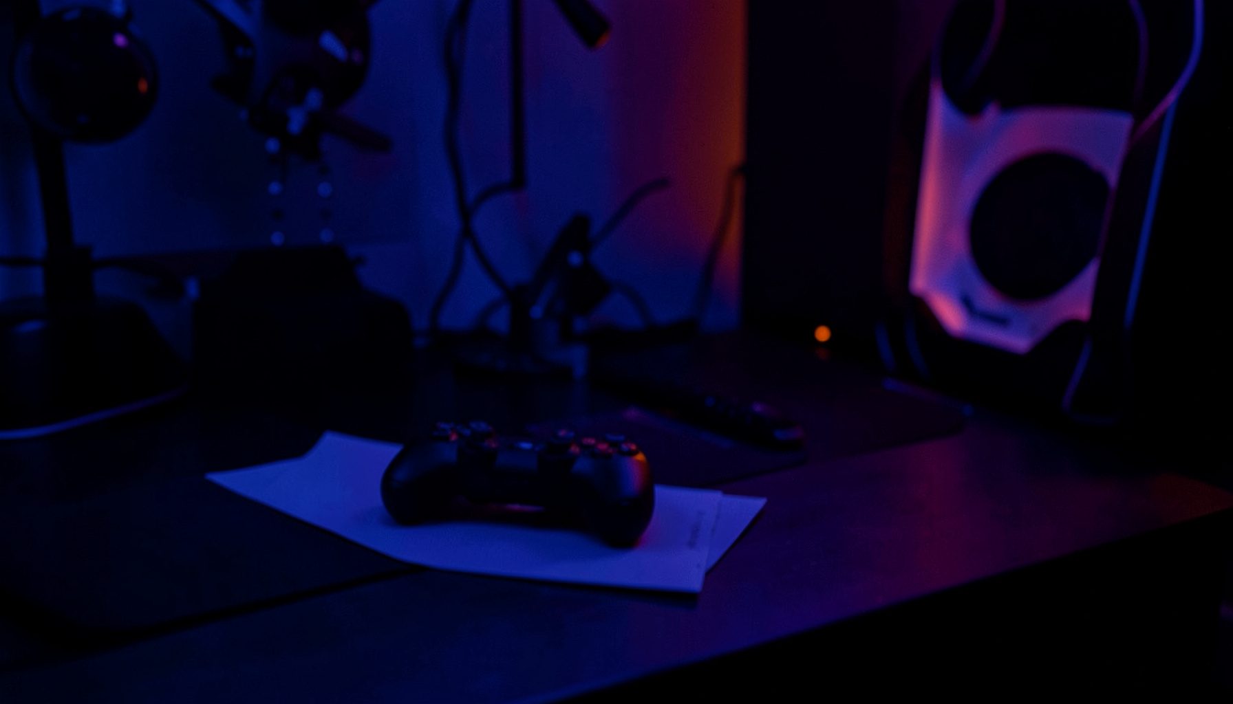 A dimly lit gaming setup with a controller resting on a desk, bathed in deep blue and violet duotone light.