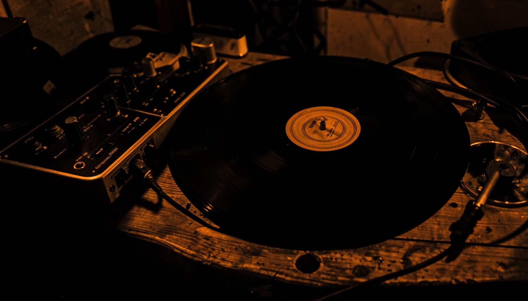 A dark atmospheric photograph of vinyl records and audio equipment on a wooden surface, deep blacks and warm amber highlights in a hard two-tone duotone