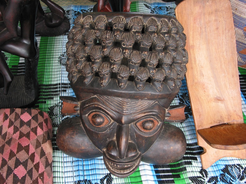 Carved wooden mask with multiple small faces displayed among patterned fabric, shown in a warm brown and cream duotone