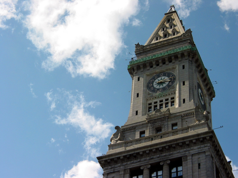 A tall stone clock tower with ornate architectural details set against a blue sky with clouds; calm daytime mood with light stone tones